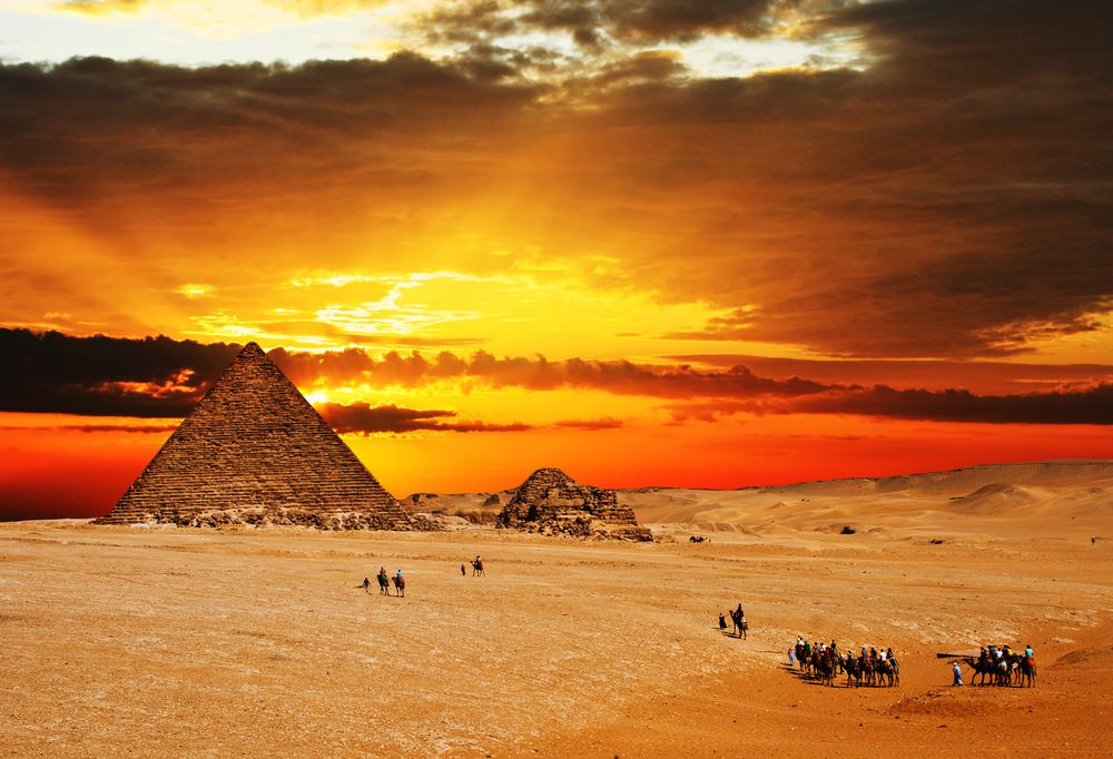Camel caravan going through desert in front of pyramid at sunset.
